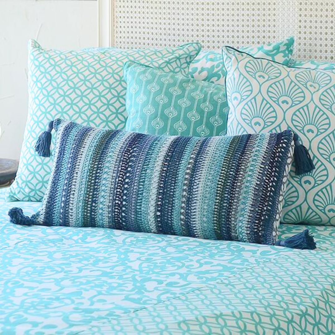 Handcrafted crochet cushion cover in blue striped pattern, displayed with coordinating teal and aqua patterned pillows on a bed, showcasing bohemian style and artisanal craftsmanship