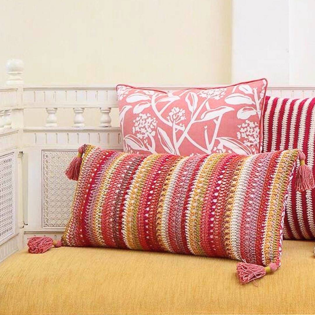 Handmade crochet cushion covers in warm confetti stripes, featuring colorful cotton twine patterns and tassels, displayed on a yellow couch with a floral pink pillow, creating a cozy boho atmosphere.