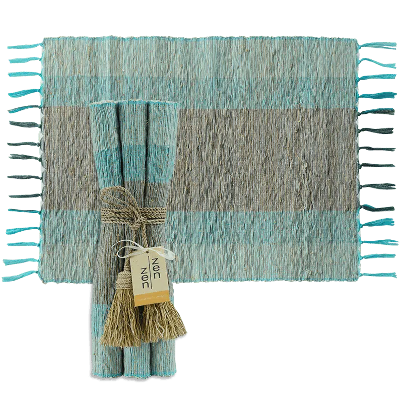 Handwoven vetiver placemats in cool grey and turquoise stripes with fringed edges, displayed alongside rolled placemats tied with natural rope and tassels, showcasing artisanal craftsmanship and natural textures