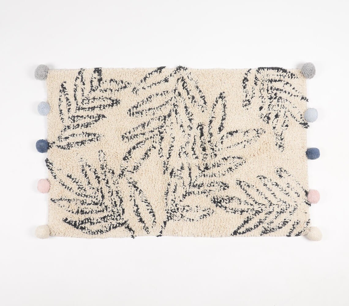 Hand-tufted cotton bath mat with leaf pattern design in black and cream, featuring decorative pom-poms along the edges in various shades of blue, gray, and pink, adding a stylish and cozy touch to bathroom decor.