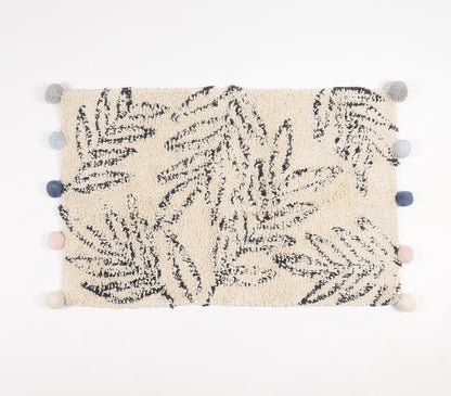 Hand-tufted cotton bath mat with leaf pattern design in black and cream, featuring decorative pom-poms along the edges in various shades of blue, gray, and pink, adding a stylish and cozy touch to bathroom decor.