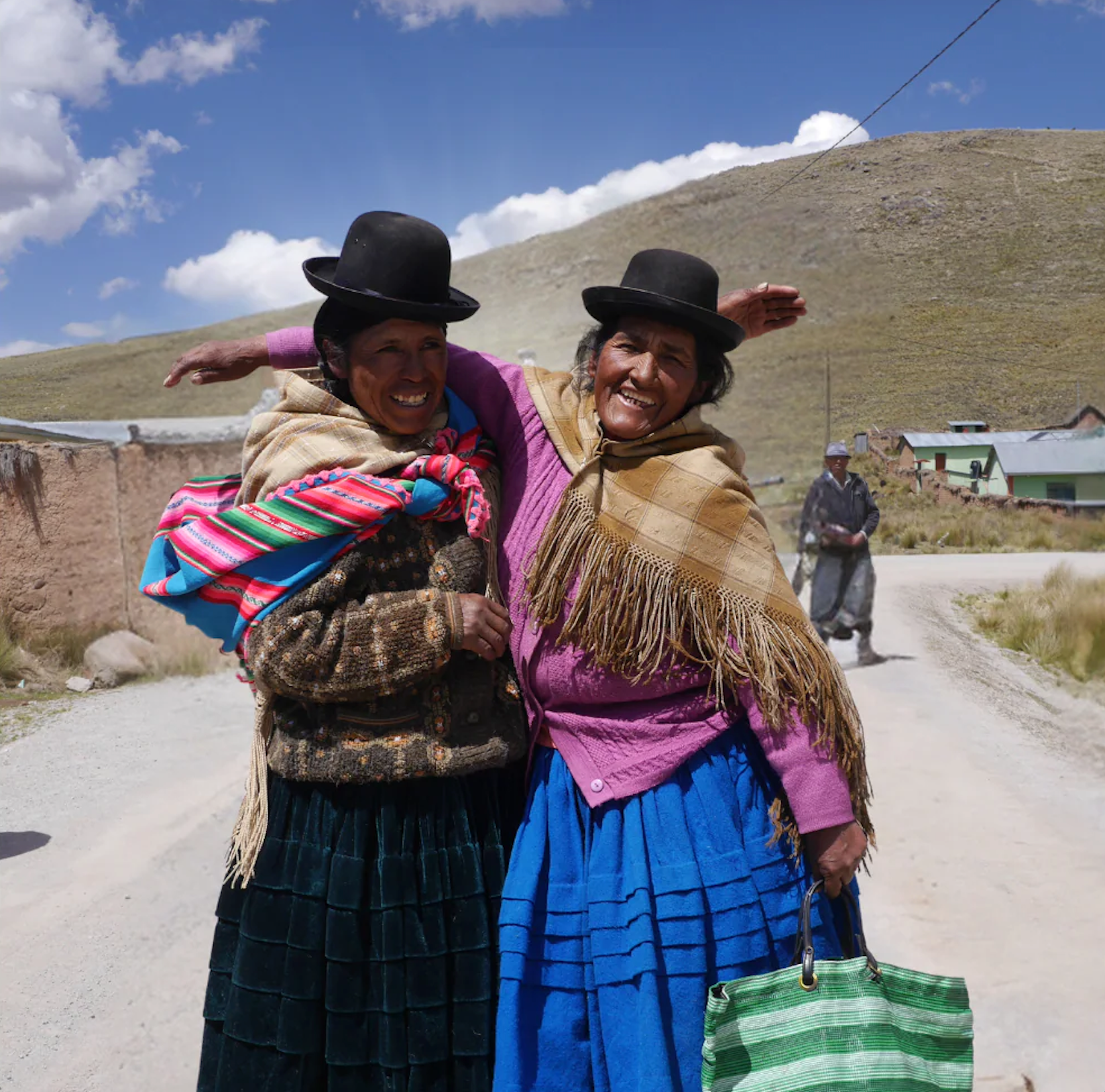 Two Peruvian women in traditional clothing standing on a dirt road with a scenic background.